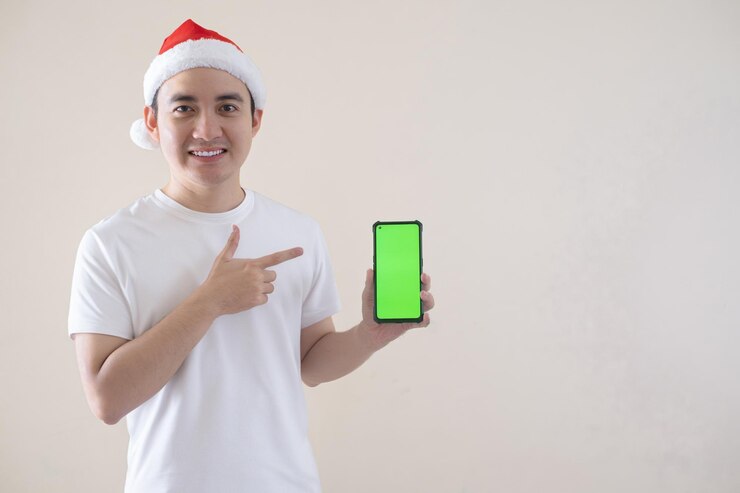 El joven Papá Noel asiático está señalando su teléfono inteligente de pantalla verde con una sonrisa