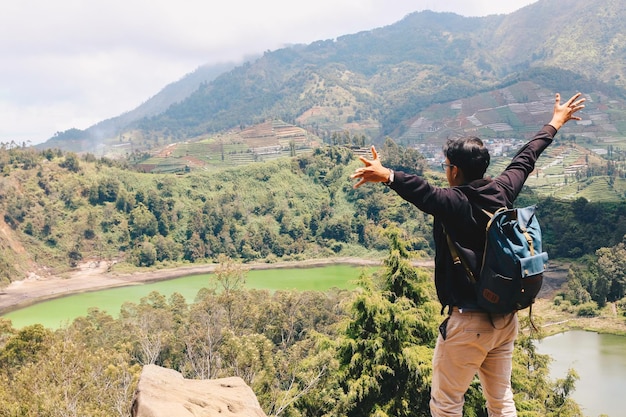 Joven parado en una roca de un acantilado y disfrutando de la vista de la naturaleza del lago y la montaña en Dieng Indonesia