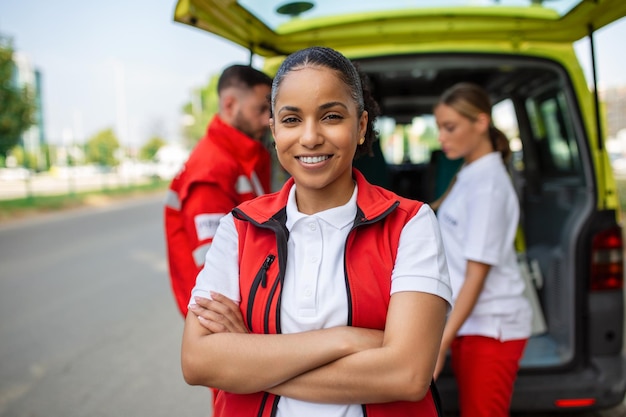 Joven paramédico afroamericano de pie detrás de los paramédicos de la ambulancia junto a la ambulancia Dos paramédicos sacando la camilla de la ambulancia