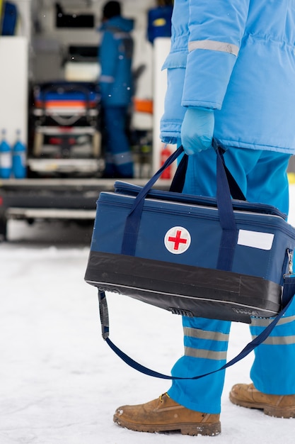 Joven paramédico en ropa de trabajo azul y guantes médicos con botiquín de primeros auxilios mientras está parado no lejos del coche de la ambulancia