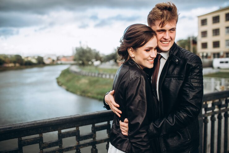 Foto joven pareja abrazados en un puente sobre un río. la novia y el novio en chaquetas de cuero. pareja amorosa en un frío día de otoño pasear por la ciudad. panorama de la ciudad con un cielo tormentoso