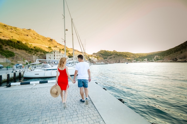 Joven pareja abrazos y relajarse en el muelle cerca del barco, en un día soleado de verano. La mujer y el hombre en ropa de moda se encuentra cerca del yate de lujo. Concepto de vida de lujo. Pareja de enamorados viajando, tiempo de luna de miel.