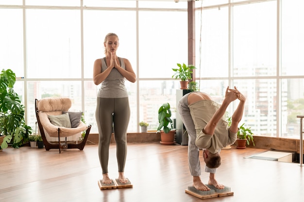 Joven pareja activa descalza en ropa deportiva de pie sobre almohadillas de terapia de yoga con cerdas metálicas mientras hacen ejercicio juntos en una habitación grande