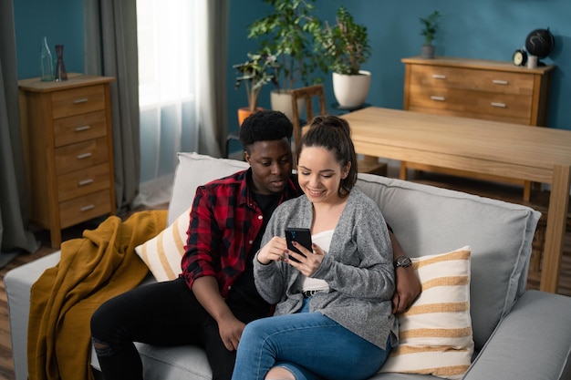 Joven pareja afroamericana alegre sentada en el sofá y viendo algo en la pantalla del teléfono inteligente