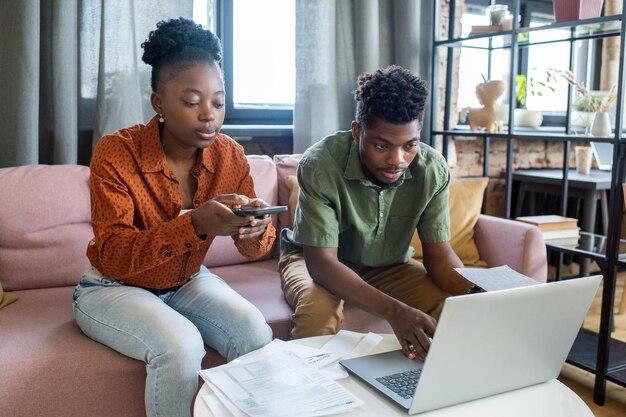 Joven pareja afroamericana en trajes casuales utilizando dispositivos modernos mientras prepara documentos fiscales