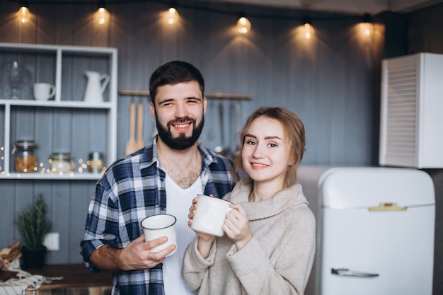 Joven pareja alegre feliz juntos sosteniendo tazas de café en la acogedora cocina.