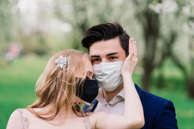 Joven pareja amorosa caminando en máscaras médicas en el parque durante la cuarentena en el día de su boda.