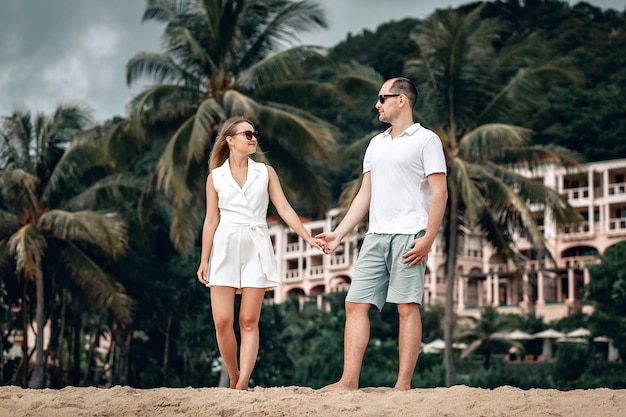 Joven pareja amorosa vestida de blanco tomados de la mano en una playa exótica sonriendo, palmeras, hotel; concepto de sentimientos.