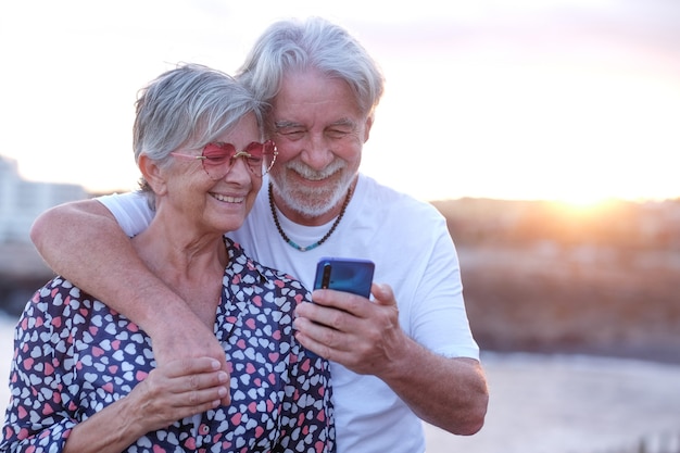 Joven pareja de ancianos abrazando al aire libre en el mar al atardecer mediante teléfono móvil personas caucásicas