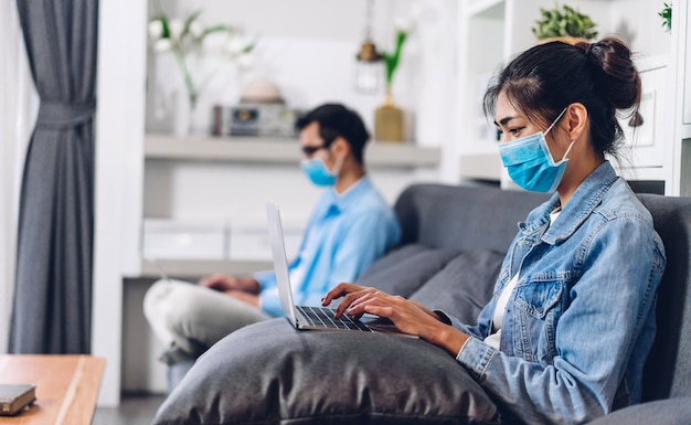 Joven pareja asiática con computadora portátil trabajando y video conferencia reunión chat en línea en cuarentena para coronavirus con máscara protectora con distanciamiento social en el hogar. trabajar desde casa concepto