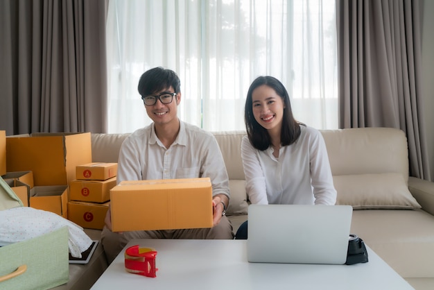 Una joven pareja asiática está vendiendo en línea a través de una computadora y ayudando a empacar la caja en la sala de estar de la casa. PYME emprendedora de pequeñas empresas o concepto independiente