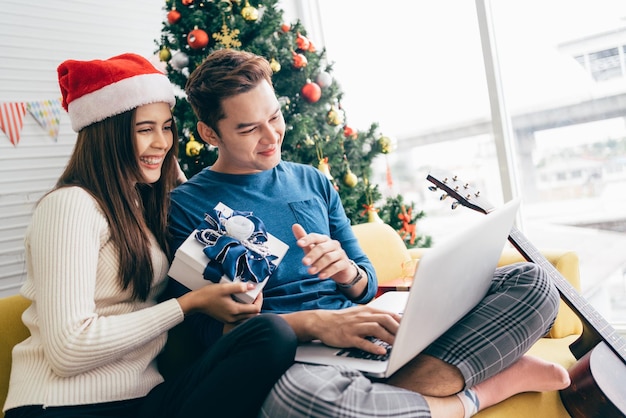 Joven pareja asiática feliz sentada juntos videollamadas en una laptop con amigos o padres gracias por un regalo recibido durante las vacaciones de Navidad con un árbol de Navidad decorado en el fondo