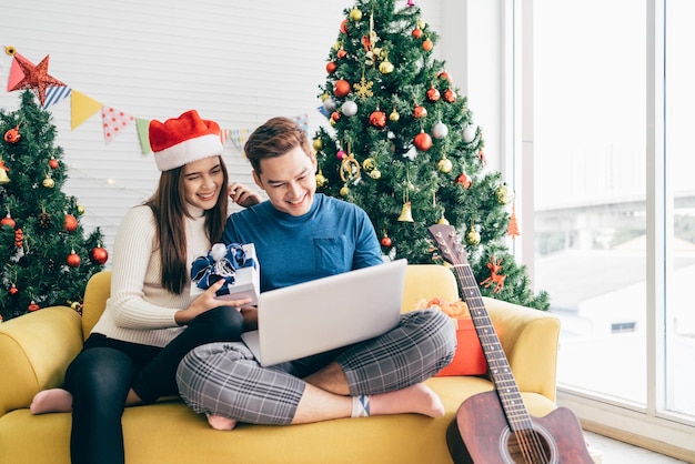 Joven pareja asiática feliz sentada juntos videollamadas en una laptop con amigos o padres gracias por un regalo recibido durante las vacaciones de Navidad con un árbol de Navidad decorado en el fondo