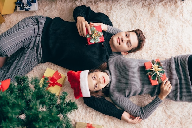 Joven pareja asiática feliz usando suéter acostado juntos y sosteniendo regalos con muchas cajas de regalo en la alfombra en la sala de estar durante las vacaciones de Navidad