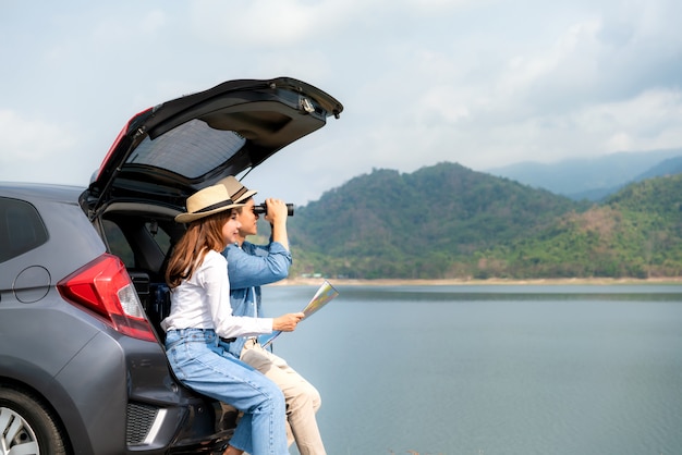 Joven pareja asiática con hombre turista mirando a través de binoculares mientras su novia sonriente sentada cerca con mapa en coche