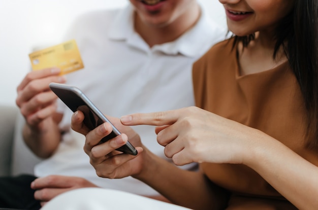Joven pareja asiática ingresando el código de seguridad en el teléfono inteligente móvil y pagando con tarjeta de crédito en el sofá en la oficina en casa