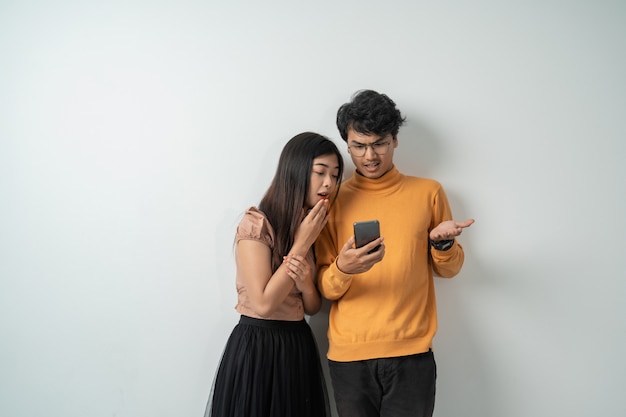 Joven pareja asiática de pie mirando la pantalla de una expresión de sorpresa de teléfono inteligente