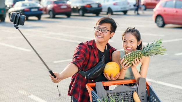 Joven pareja asiática sosteniendo en sus manos piña y melón y haciendo selfie foto.