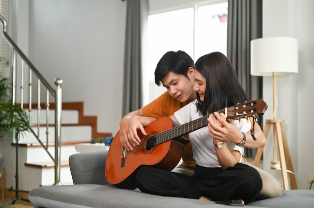 Joven pareja asiática toca la guitarra y canta una canción en el sofá en casa juntos.