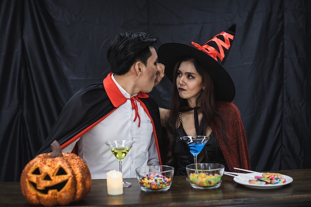 Joven pareja asiática en traje de bruja y Drácula con celebrar la fiesta de Halloween para tintinear el vidrio y beber en el festival de Halloween. Pareja en traje celebra la fiesta de Halloween de fondo de tela negra.