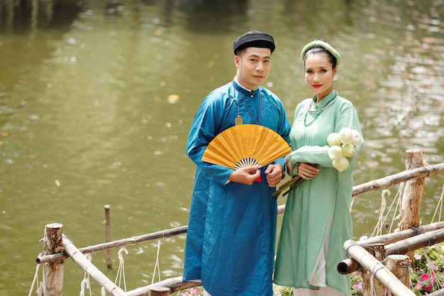 Joven pareja asiática en trajes tradicionales de pie en el estanque con flores de loto y abanico de papel y mirando a la cámara