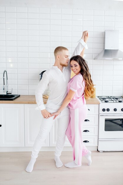 Joven pareja bailando en la cocina, linda chica europea con el pelo largo en traje rosa casero, moderna cocina blanca