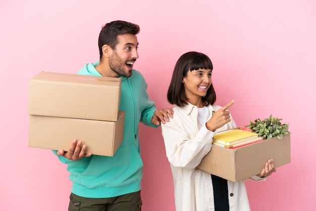 Joven pareja de belleza mudándose a un nuevo hogar entre cajas aisladas en un fondo rosa apuntando hacia un lado para presentar un producto