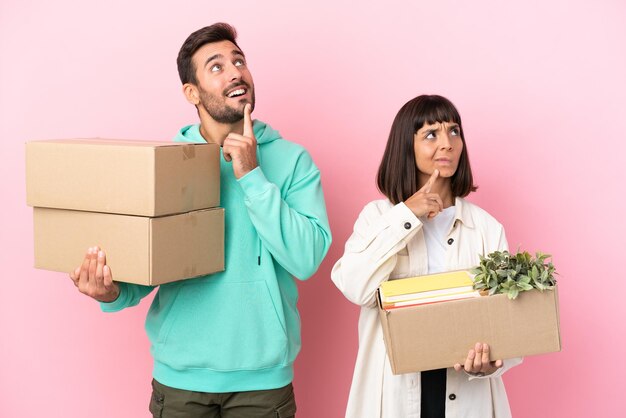 Joven pareja de belleza mudándose a un nuevo hogar entre cajas aisladas en un fondo rosa pensando en una idea mientras mira hacia arriba