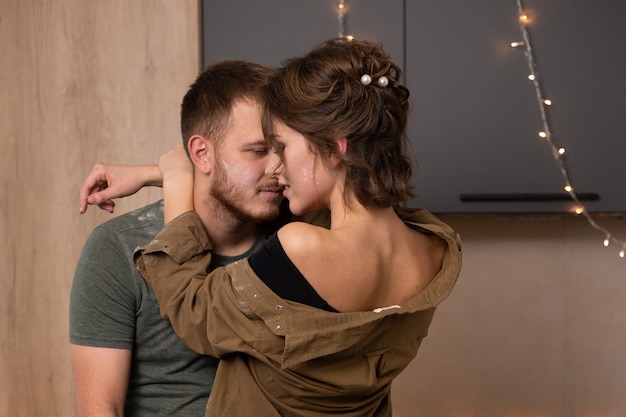 Joven pareja besándose y sonriendo mientras cocina en la cocina de su casa.