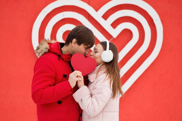 Joven pareja besándose y sosteniendo corazón de papel cerca de gran corazón en la pared.