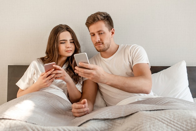 Joven pareja en la cama. Una mujer y un hombre atractivos están revisando juntos las noticias de las redes sociales en los teléfonos móviles, chateando, enviando mensajes de texto o enviando mensajes