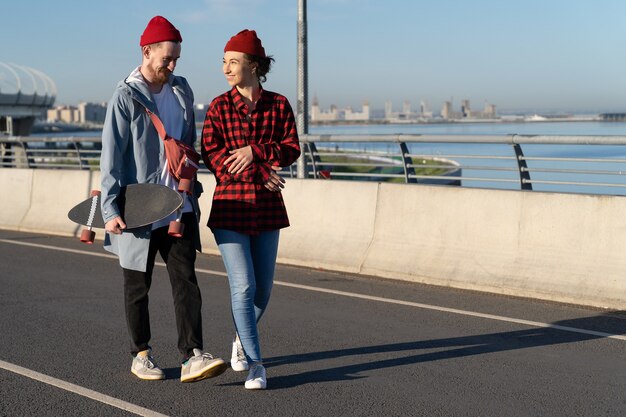 Joven pareja casual de hipsters camina por la calle de la ciudad con charla de longboard sobre el fondo del horizonte urbano