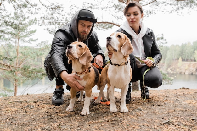 Joven pareja casual sentada en cuclillas abrazando a dos cachorros beagle de pura raza mientras se relaja en un entorno rural