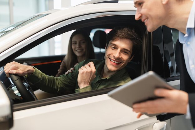 Joven pareja caucásica aclarando los detalles de la compra de automóviles con un vendedor mirando la pantalla de la tableta en