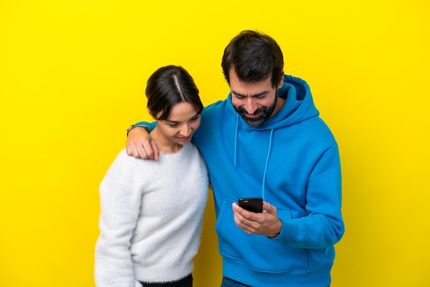 Joven pareja caucásica aislada de fondo amarillo enviando un mensaje o correo electrónico con el móvil