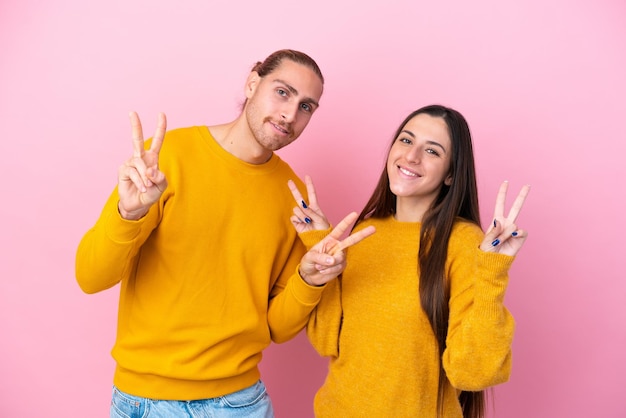 Joven pareja caucásica aislada de fondo rosa sonriendo y mostrando el signo de la victoria con ambas manos