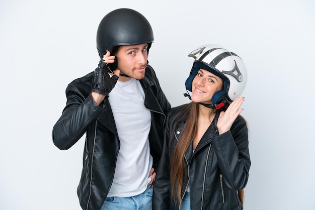 Foto joven pareja caucásica con un casco de motocicleta aislado de fondo blanco escuchando algo poniendo la mano en la oreja