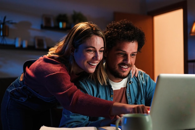 Joven pareja caucásica feliz navegando en Internet usando una computadora portátil para buscar venta en casa Marido emocionado y esposa alegre sonriendo y buscando un nuevo apartamento sentado en un escritorio en la sala de estar