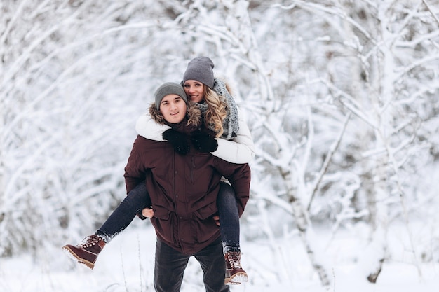 Joven pareja caucásica hombre y mujer cabalgando sobre sus espaldas en vacaciones de invierno en un bosque nevado