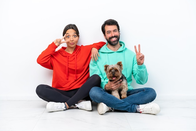 Joven pareja caucásica sentada en el suelo con su mascota aislada de fondo blanco sonriendo y mostrando el signo de la victoria