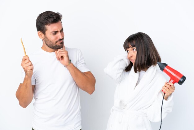 Joven pareja caucásica sosteniendo un secador de pelo y un cepillo de dientes aislado de fondo blanco que tiene dudas mientras se rasca la cabeza