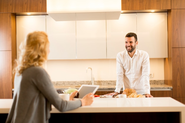 Joven pareja en la cocina, una mujer sentada con una tableta en la mano mientras el hombre prepara la comida