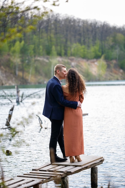 Una joven pareja cogidos de la mano sobre un puente de madera en medio de un lago azul. Albañilería en la isla sobre un fondo de árboles. Naturaleza, paisaje. Romance y amor, pareja feliz