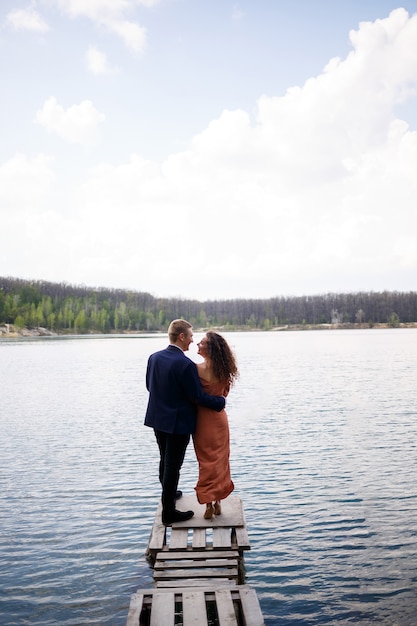 Una joven pareja cogidos de la mano sobre un puente de madera en medio de un lago azul. Albañilería en la isla sobre un fondo de árboles. Naturaleza, paisaje. Romance y amor, pareja feliz