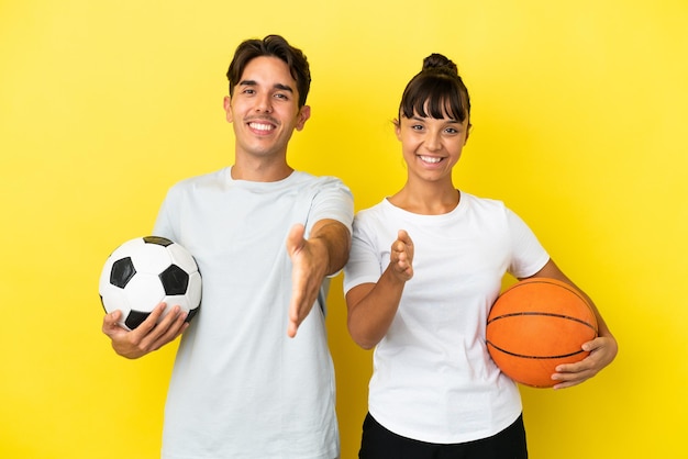 Joven pareja deportiva jugando fútbol y baloncesto aislada en un fondo amarillo estrechando la mano para cerrar un buen trato