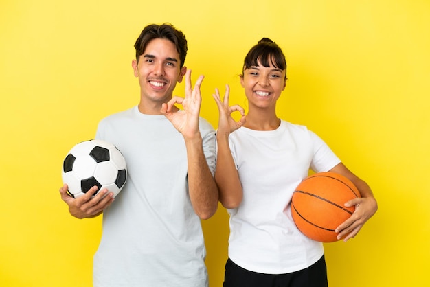 Joven pareja deportiva jugando fútbol y baloncesto aislada en un fondo amarillo mostrando un signo de ok con los dedos