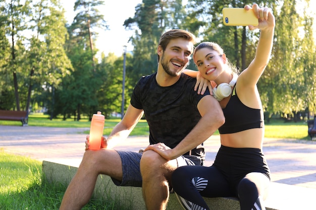 Joven pareja deportiva en ropa deportiva sentada en el parapeto en el parque urbano y tomando selfie después del entrenamiento en un día soleado de verano.