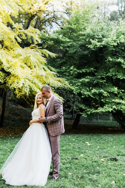 Joven pareja disfrutando de momentos románticos mientras camina en el parque. Elegante novia y el novio posando y besándose en el parque el día de su boda. Elegante novia en un hermoso vestido blanco, el novio en un traje.