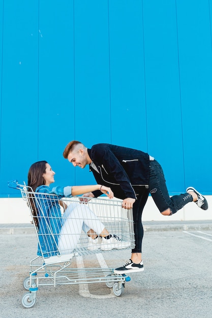 Joven y pareja divirtiéndose en un carrito de compras en fondo azul.