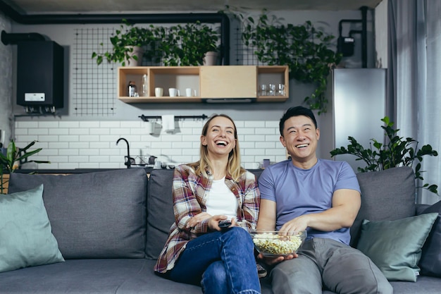 Joven pareja enamorada de hombre y mujer viendo la televisión juntos en casa regocijándose y comiendo palomitas de maíz feliz familia multirracial en un apartamento nuevo